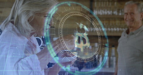 Tapping senior woman and smiling senior man examining glowing Bitcoin interface at wooden counter