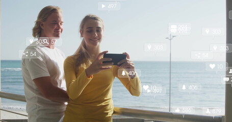 Snapping selfie couple wearing mustard top and white tee standing on seaside deck with smartphone