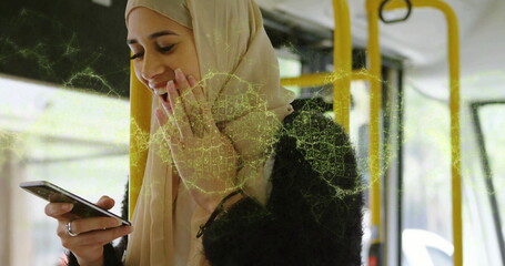 Smiling woman wearing hijab and black jacket, holding phone inside bus by windows with yellow poles