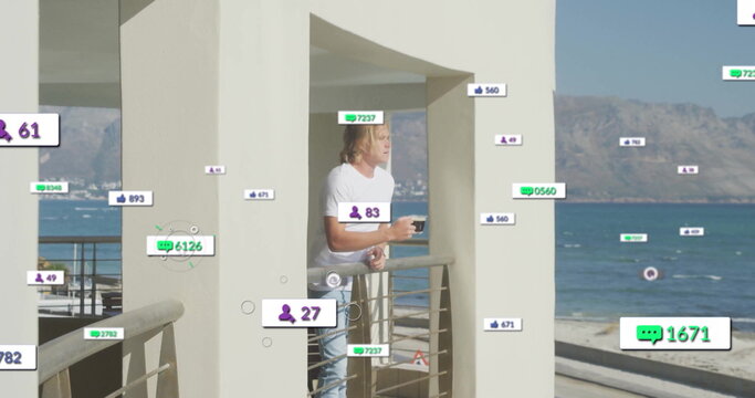 Gazing man wearing t-shirt and jeans holding coffee mug on seaside balcony, with engagement icons