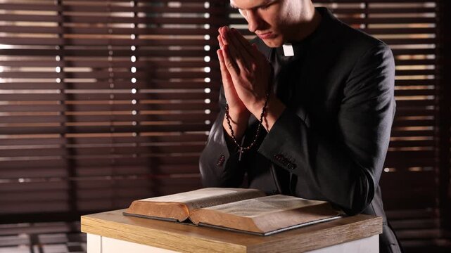 Priest with Bible and rosary praying at table in confessional booth, closeup