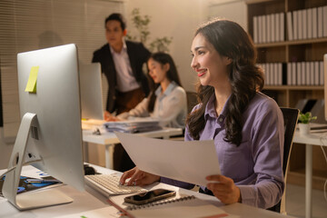 A woman is sitting at a desk with a computer monitor and a stack of papers