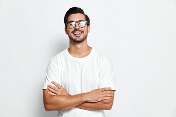 Happy smiling young man with glasses wearing a casual white T-shirt, standing confidently with crossed arms against a plain solid background. Personal branding concept.