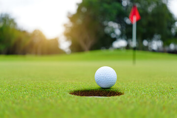A golf ball sits on the green grass of a golf course near a hole with a red flag in the background, ready for the next shot.