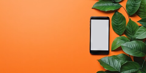 Flat lay of a black smartphone with a blank screen surrounded by vibrant green leaves on an orange background