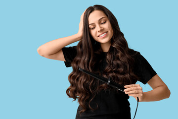Beautiful young woman with curling iron on blue background