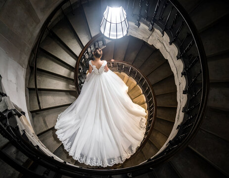 A radiant bride descends a dramatic spiral staircase, her exquisite wedding gown elegantly cascading under the soft glow of a top spotlight, creating a timeless and ethereal scene. - Powered by Adobe
