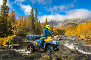 Man on quad bike crosses river. Guy on ATV travels through nature. Quad biker in coniferous autumn forest. Man on quad bike stands in middle of turbulent river. Quad biker in helmet looks around © Grispb