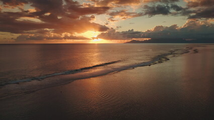 Golden sunset creates a magical atmosphere over the calm ocean waves gently lapping against the shore in Tahiti, French Polynesia, with distant mountains adding to the serene beauty