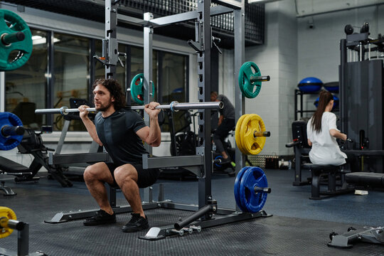 Wide shot of young Caucasian man and unrecognizable woman doing strengthening exercises with use of fitness equipment in modern gym