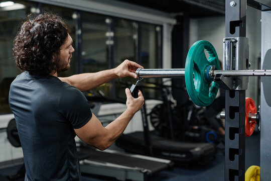 Medium shot of young Caucasian man changing weights on barbell during workout in modern gym, copy space