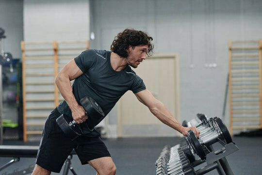 Side view of muscular young Caucasian man with curly hair exercising in modern gym using dumbbells, copy space