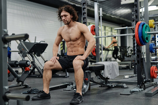 Young Caucasian man with curly hair sitting shirtless on bench in gym having break, copy space