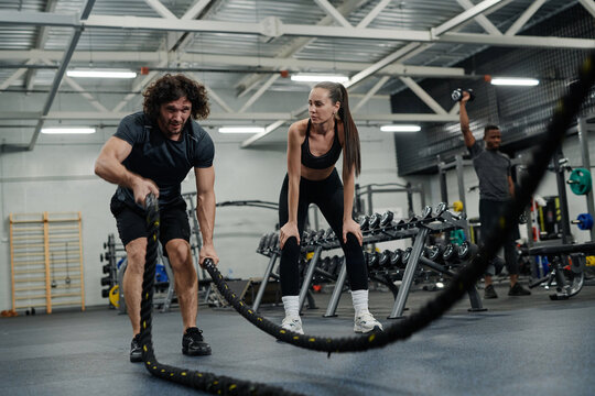 Wide shot of young woman watching strong young man doing rope wave exercise in modern gym, copy space