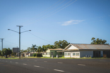 A long row of modest, low-slung buildings with corrugated metal roofs lines a quiet street under a clear, blue sky with high, thin clouds. A streetlight stands in the foreground. West QLD, Australia. © pauline.mongarny