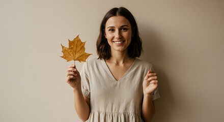 Smiling Woman Holding Autumn Leaf: Portrait of a happy young woman in beige dress holding a dry maple leaf against a neutral ba...