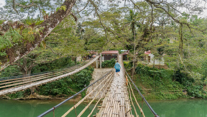 A man walks on a rickety bamboo suspension bridge over a river. Flimsy handrails and woven walkways. Tropical vegetation on the shore. Philippines. Bohol. Bamboo Hanging Bridge. Sipatan River  