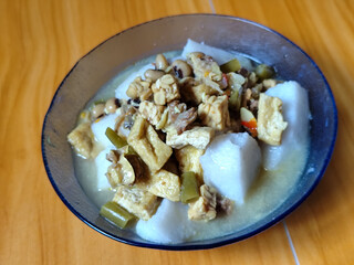 A bowl of vegetable rice cakes. Lontong vegetable in coconut milk sauce with tofu, beef which is a typical Indonesian food. Served on a wooden table.