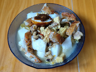 A bowl of vegetable rice cake. Lontong vegetable with coconut milk sauce and accompaniments of Indonesian tofu, beef and petis eggs, served on a wooden table.