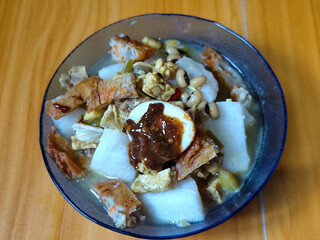 A bowl of vegetable rice cake. Lontong vegetable with coconut milk sauce and accompaniments of Indonesian tofu, beef and petis eggs, served on a wooden table.