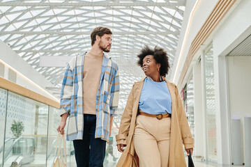 Horizontal low angle view shot of young Caucasian man and Black woman talking while walking along mall