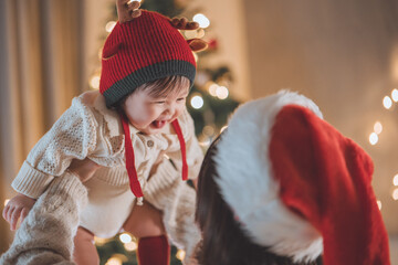 Asian mother wearing Santa hat holding cheerful little daughter in red reindeer hat indoors near decorated Christmas tree and lights, celebrating baby’s first Christmas in warm festive holiday season