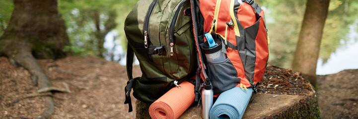 Header of two backpacks with attached yoga mats and water bottles resting on tree stump near forest stream, suggesting outdoor adventure or camping activity, no people visible