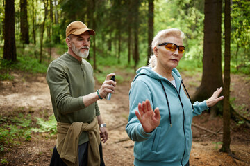 Senior Caucasian woman walking in forest raising hand while senior Caucasian man following behind holding insect repellent spray preparing to apply it outdoors