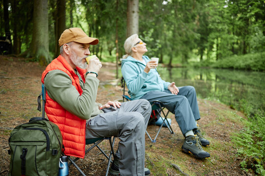 Senior Caucasian man eating sandwich and senior Caucasian woman drinking from cup while sitting on folding chairs near forest lake, both relaxing outdoors surrounded by trees