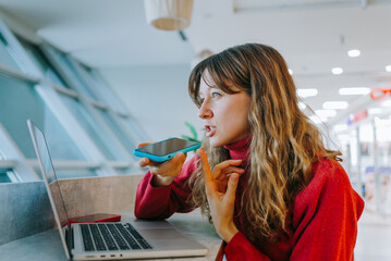 Young woman recording voice message on smartphone while working on laptop in coworking space,...