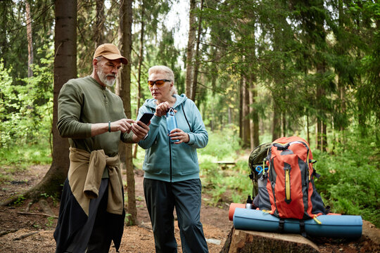 Caucasian senior man showing smartphone to Caucasian senior woman while standing in forest, both wearing outdoor clothing, large hiking backpack and camping gear visible nearby
