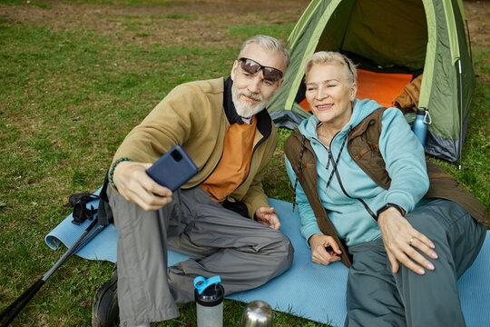 Senior Caucasian man and woman sitting on camping mats in front of tent taking selfie with smartphone, both smiling and looking at device, outdoor setting visible