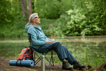 Obraz premium Senior Caucasian woman sitting on folding chair near forest lake holding cup, wearing sunglasses, relaxing outdoors with backpack and yoga mat beside, enjoying peaceful nature surroundings