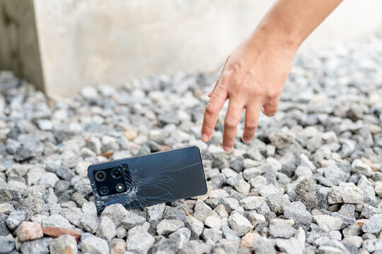 A hand is reaching down to pick up a broken cell phone. The phone is cracked and has a black screen