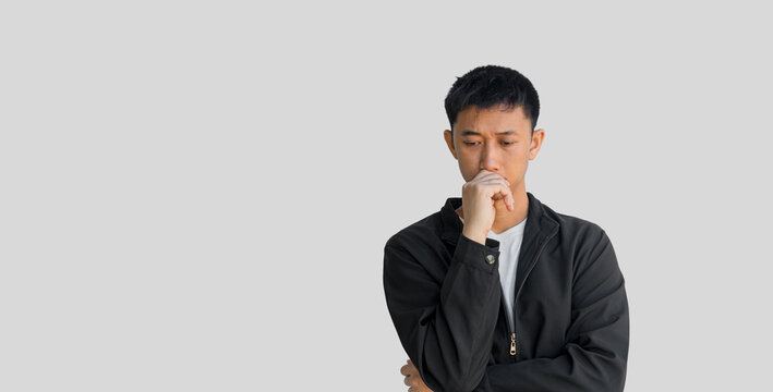 A young Asian man in his 20s wearing a black coat is looking suspicious and has a confused expression or is thinking, isolated on a grey background