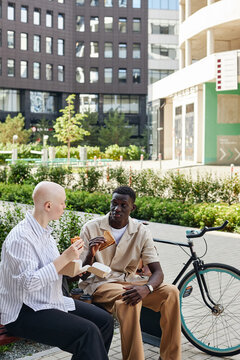 Vertical wide shot of two ethnically diverse man and woman wearing smart casual clothes sitting on bench outdoors eating lunch and chatting