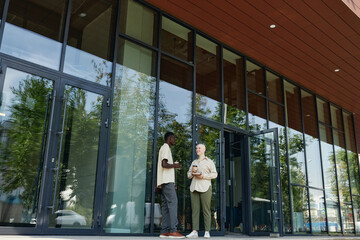 Extreme long shot of two ethnically diverse millennials standing by office building doors chatting during coffee break, copy space