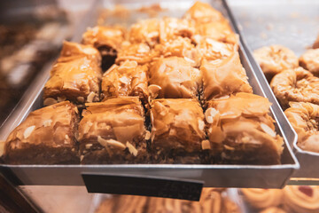 A close-up shot of square-cut, golden-brown baklava with a shiny honey glaze and scattered almond slivers, displayed on a metal tray in a bakery.