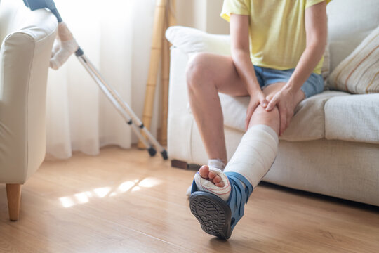 Patient wearing an orthopedic boot, with crutches nearby, massaging their injured leg while sitting on a sofa, recovering from a metatarsal fracture
