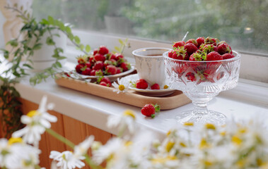 A collection of vibrant strawberries is displayed in a glass bowl on a clean windowsill, complemented by a cup of tea and cheerful daisies, creating a charming kitchen atmosphere