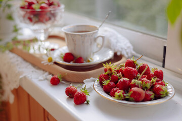 Sunlight streams through the window onto a wooden sill adorned with ripe strawberries and a cup of coffee. A tray holds more strawberries, creating a warm and inviting summer atmosphere