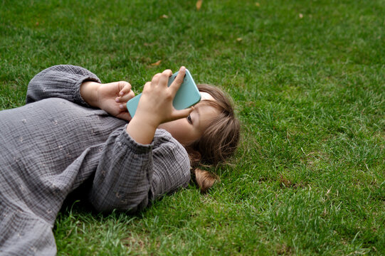 Child girl lying on grass with smartphone in garden.