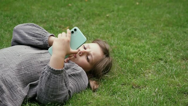 Child girl lying on the grass in a garden, using a smartphone.
