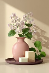 Pale pink vase holds delicate light purple flowers;  two pale cubes and eucalyptus sprig rest on a mauve tray, bathed in soft sunlight against a beige background