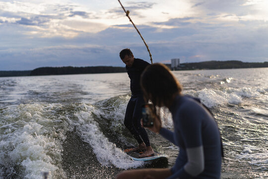Man wake surfing on lake while woman records him at sunset