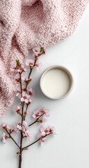 Flatlay of a pale pink knit throw, delicate pink blossoms, and a small bowl of creamy liquid on a white background