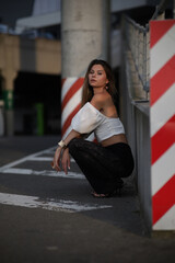 Fototapeta premium Stylish young woman in elegant black lace pants and a white off-shoulder top posing outdoors, leaning on a railing with an urban background in warm evening light.
