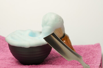 Shaving foam overflowing from a bowl with a straight razor and shaving brush resting on a pink towel, ready for a traditional wet shave
