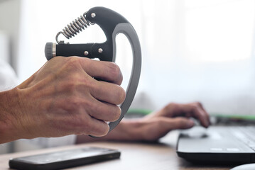Male office worker exercising with hand gripper to strengthen wrists and forearms. Man using hand grip strengthener while working on laptop at desk