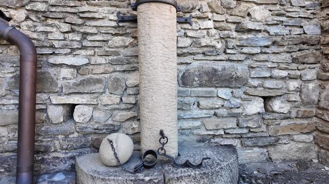 White stone pillory with black iron shackles mounted to castle walls. A heavy chained sphere lies below. Historic Slovak setting evoking medieval justice, punishment, and architectural heritage.